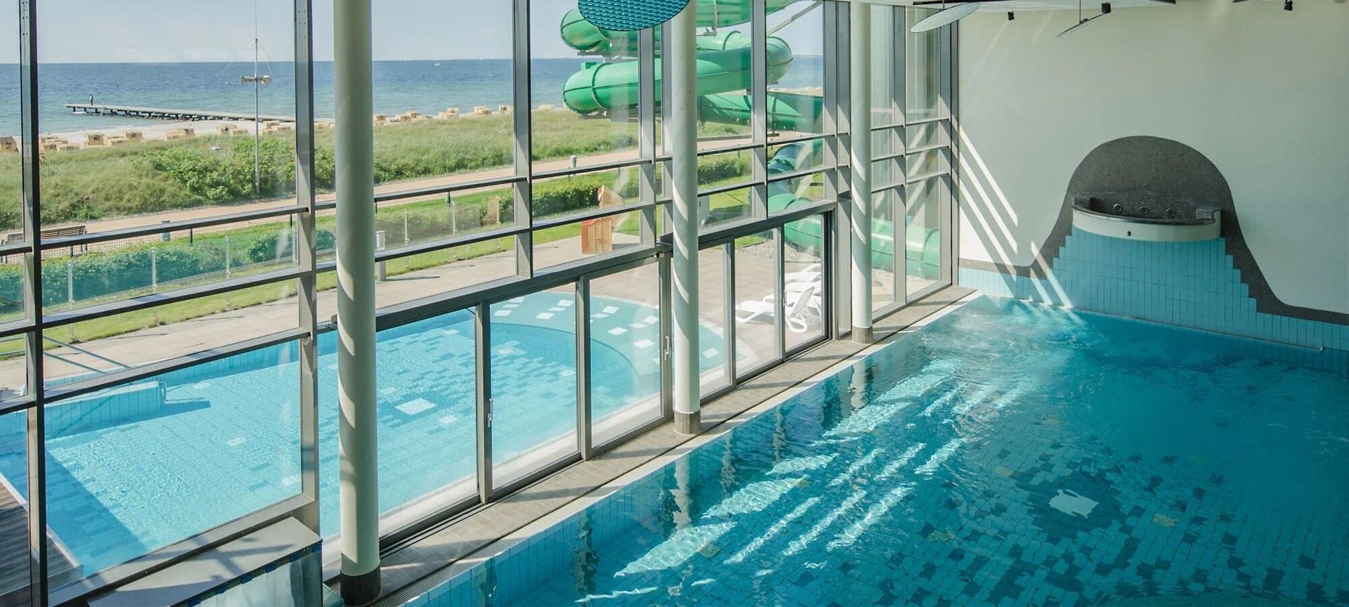 Innenansicht eines Schwimmbads mit großen Fenstern, Blick auf Außenpool, grüne Wasserrutsche und Meer im Hintergrund