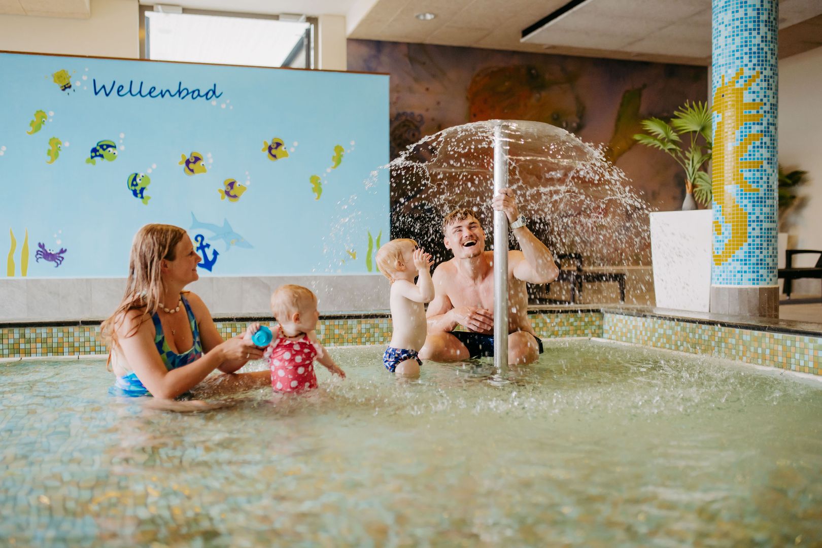 Familie mit zwei kleinen Kindern spielt im flachen Wasser eines Hallenbads unter einer Wasserfontäne