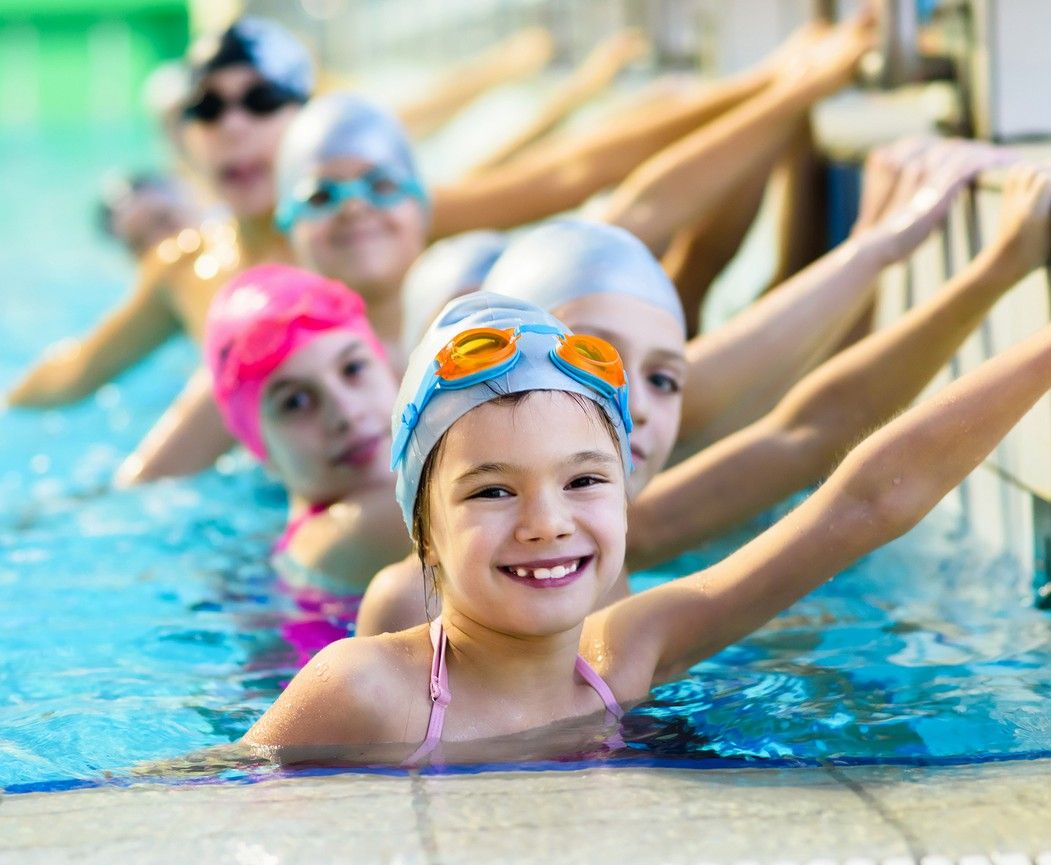 Kinder im Schwimmbecken halten sich am Beckenrand fest, tragen Schwimmkappen und Schwimmbrillen
